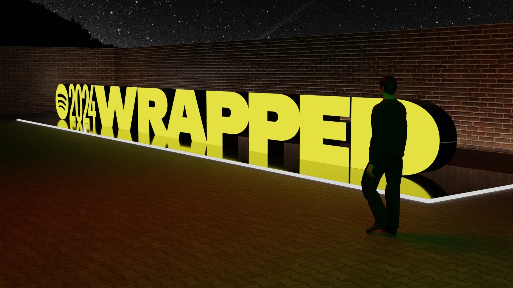 A person standing beside a large illuminated yellow 2024 Wrapped sign in front of a brick wall at night.